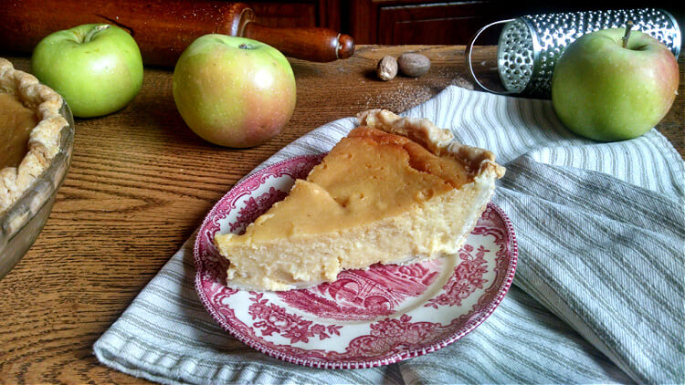 Close up of a slice of marlborough pudding on a red decorative plate with apples, a rolling pin and spice grater