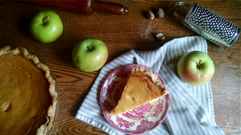 Slice of marlborough pudding on a red decorative plate with apples and the rest of the pie on a table with nutmegs and a spice grater